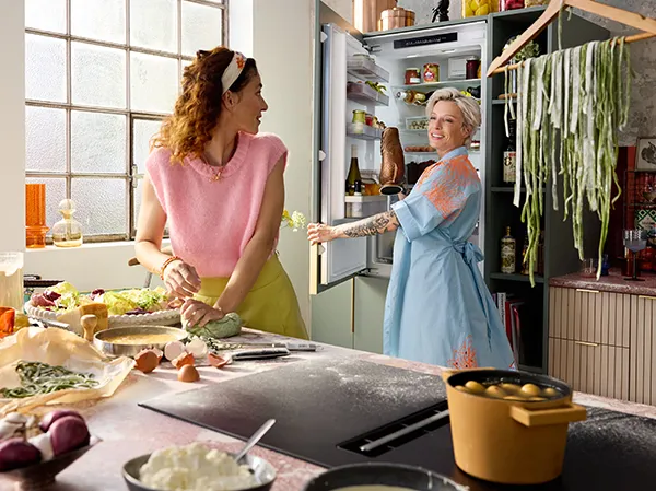 Two women in a bright kitchen prepare food together, surrounded by fresh ingredients and cooking tools, as a refrigerator stands open.
