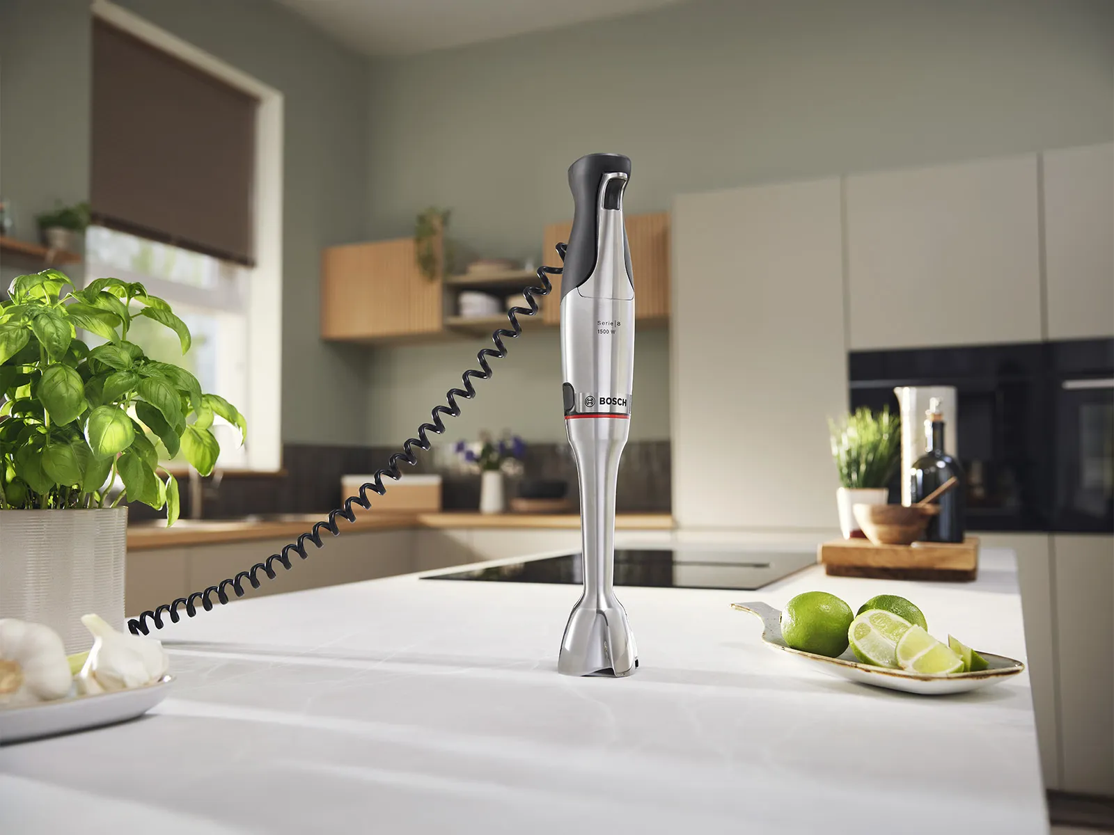 Een slanke Bosch-staafmixer staat op het keukenaanrecht, omringd door verse basilicum, knoflook en limoenen in een moderne keukenomgeving.
