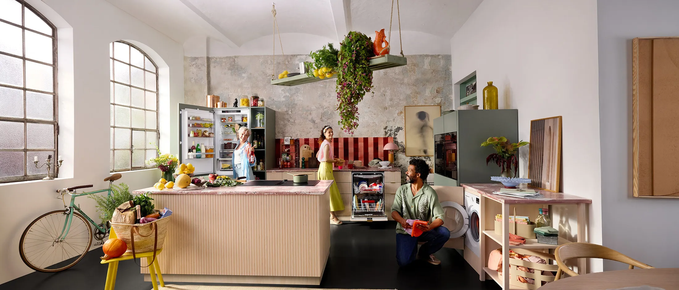 Una cucina moderna piena di prodotti freschi, dotata di un'isola, frigorifero aperto e tre persone impegnate in attività culinarie.