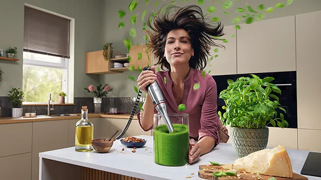 Una mujer en la cocina mezclando un batido verde con una batidora de mano, mientras las hojas de albahaca vuelan alrededor.