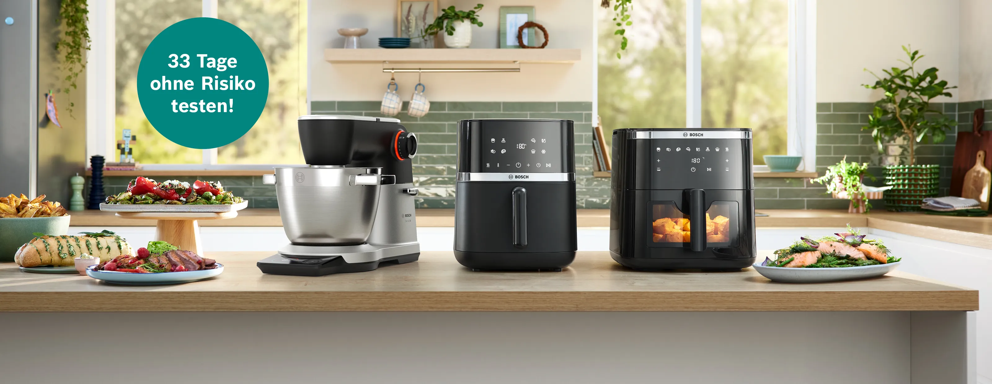 Bosch-Küchenmaschine und zwei Heißluftfritteusen auf Küchentheke, daneben zubereitete Speisen.
