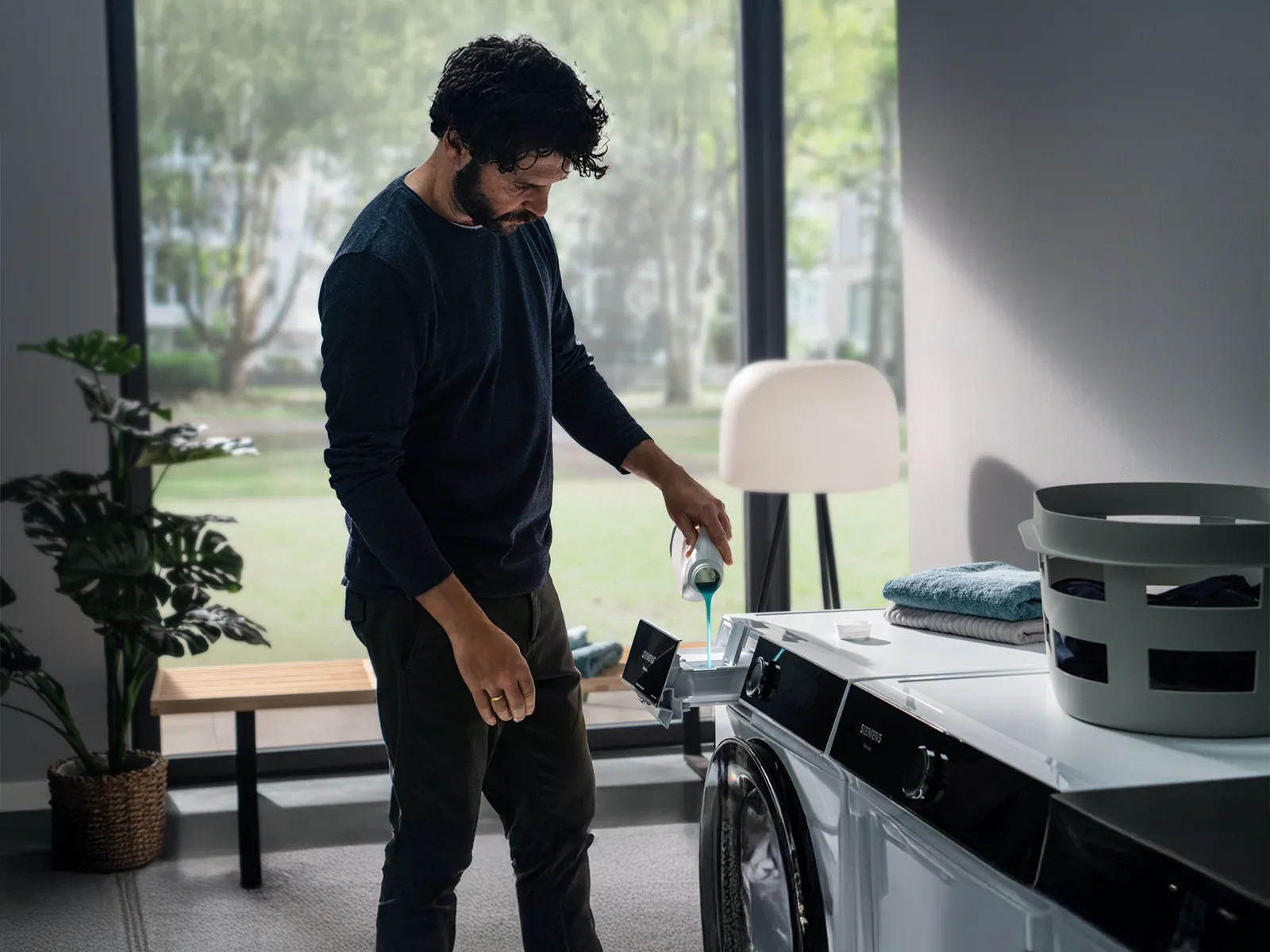 Man in black shirt standing in room with plants and furniture.