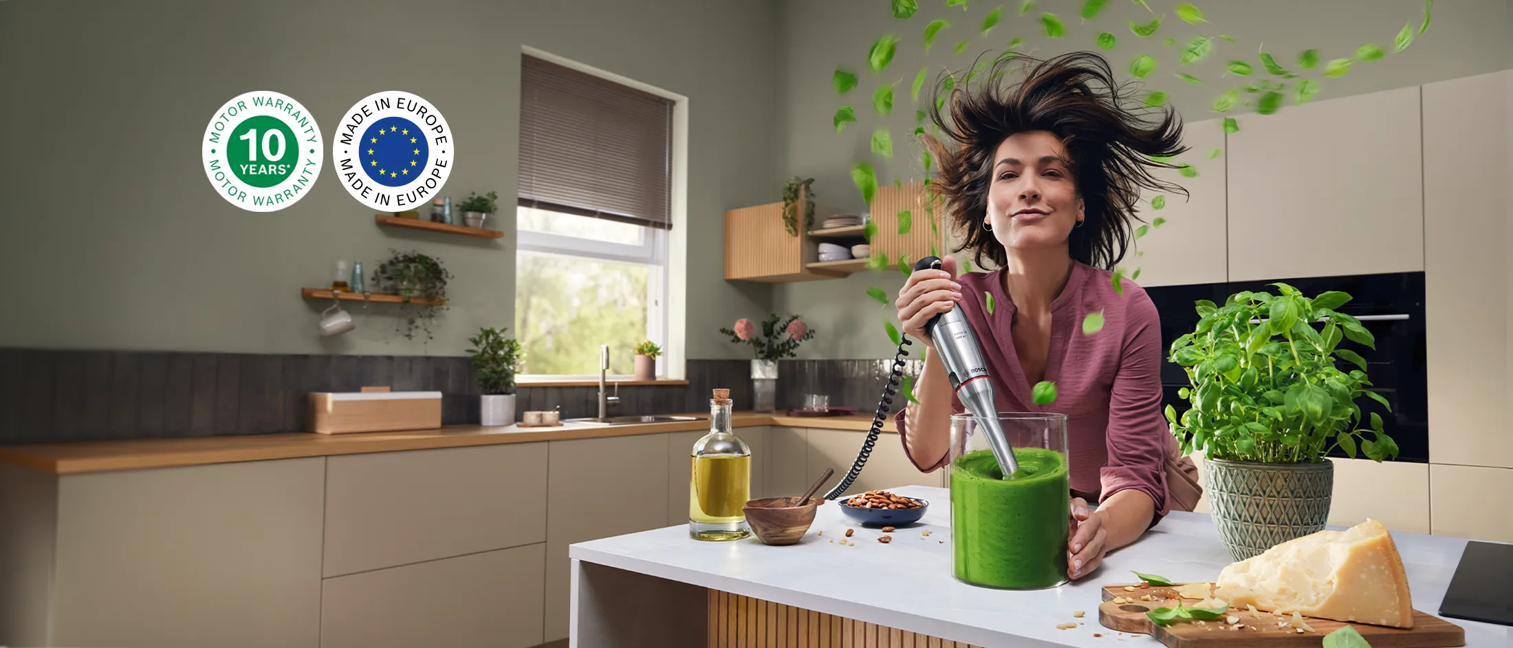 Smilende kvinne blender en grønn smoothie på et lyst kjøkken, med friske blader som flyr, omgitt av nøtter, ost og basilikum.