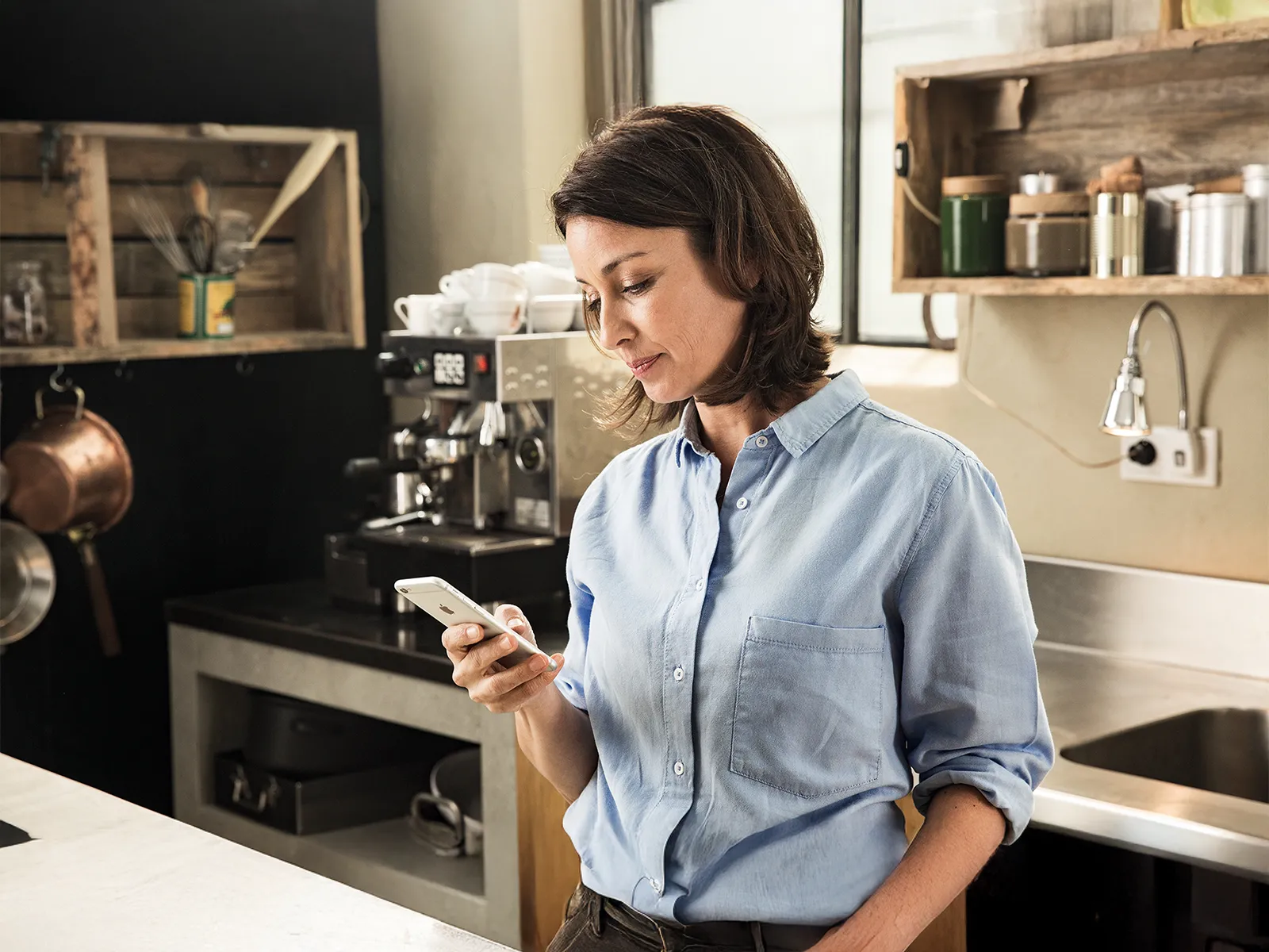 A woman in a light blue shirt stands in a stylish kitchen, holding a smartphone and looking down at the screen.