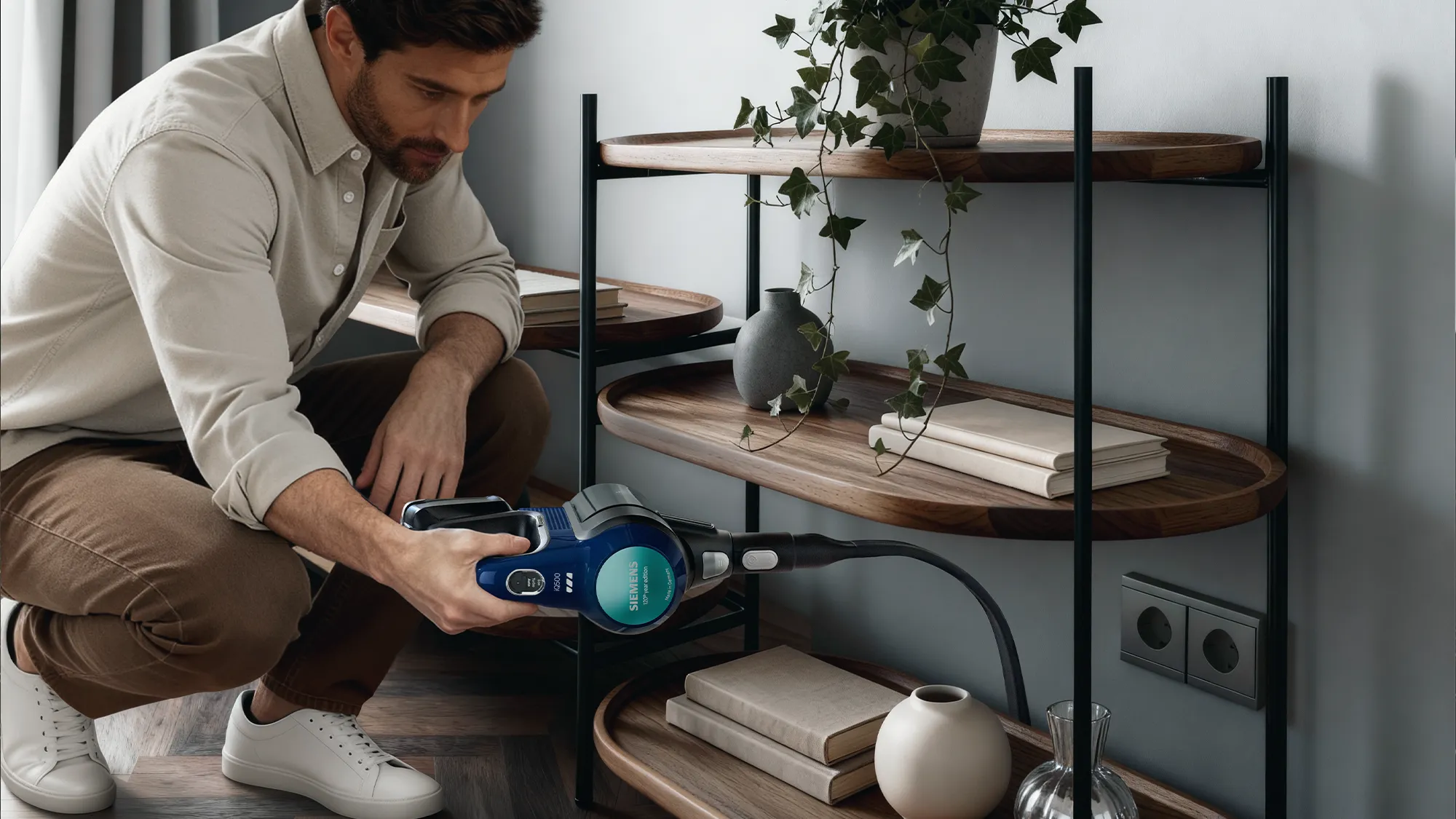 A man crouching to plug in a blue Siemens handheld vacuum cleaner to a wall outlet next to a wooden shelf.