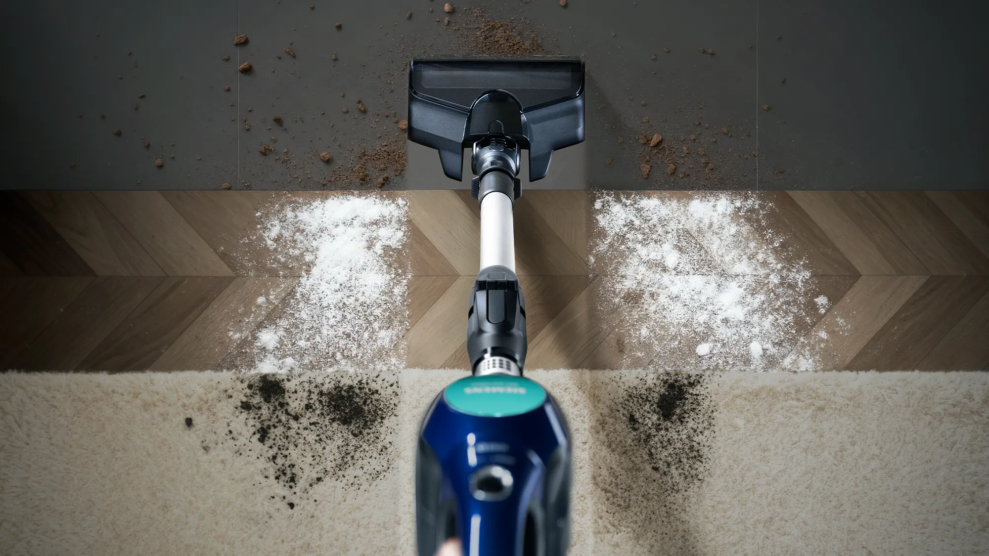A vacuum cleaner head on a floor transitioning from tile to wood to carpet, with dirt and dust showing its cleaning path.