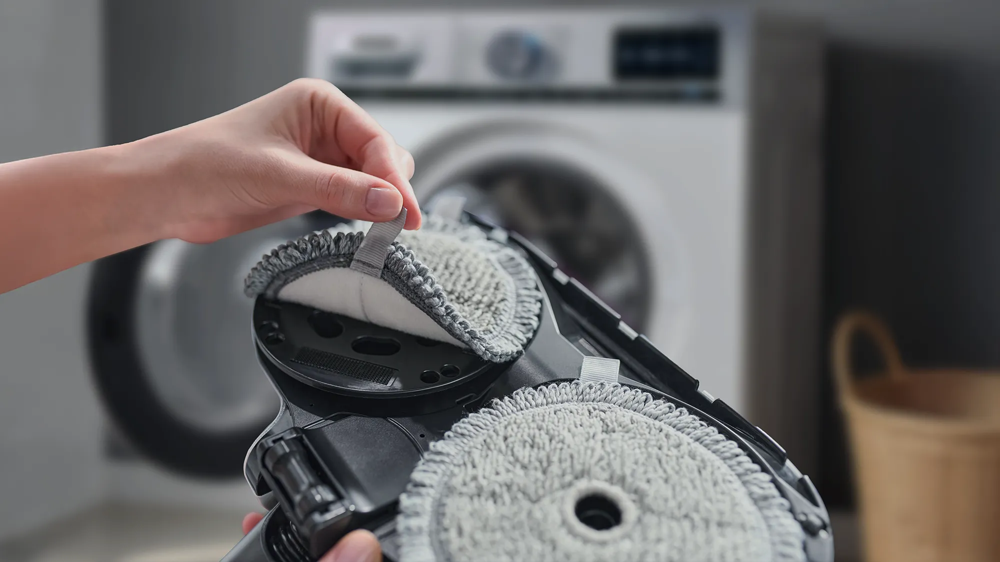 A hand removing a grey cleaning pad from a floor cleaning device, with a washing machine in the background.