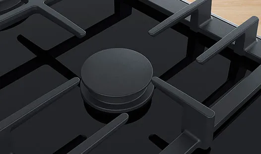 Mustan keraamisen kaasukeittotason kulma, jossa näkyvät poltin ja kattilankannattimet.
