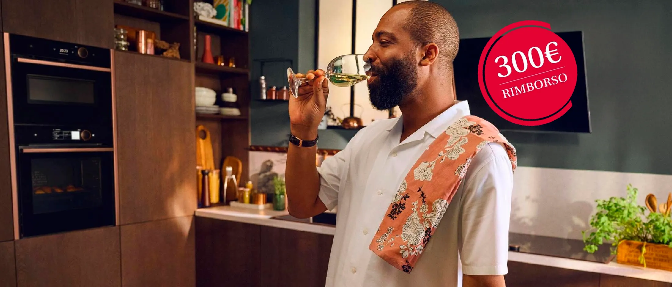 Una persona in una cucina moderna, tiene un bicchiere e indossa una camicia bianca con un asciugamano colorato sulle spalle. Sulla destra il logo "Cashback 300€"