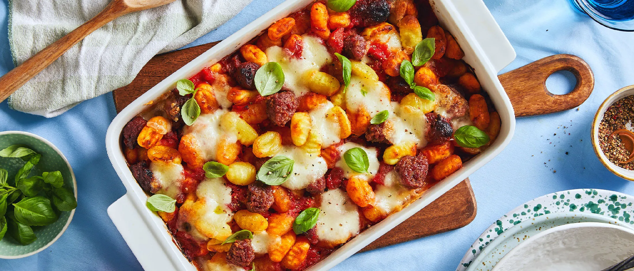 Бяла тава за печене, пълна с ньоки и наденица в доматен сос, стои на светлосиня покривка и е заобиколена от други съдове за хранене.