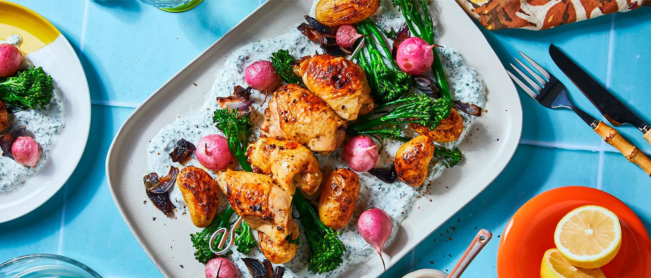 A plate of air fried vegetables and chicken breasts on a herby yogurt dip on a blue underground surrounded by colorful dinner table dishes, like a fork, a napkin, plates and a glas filled with water.