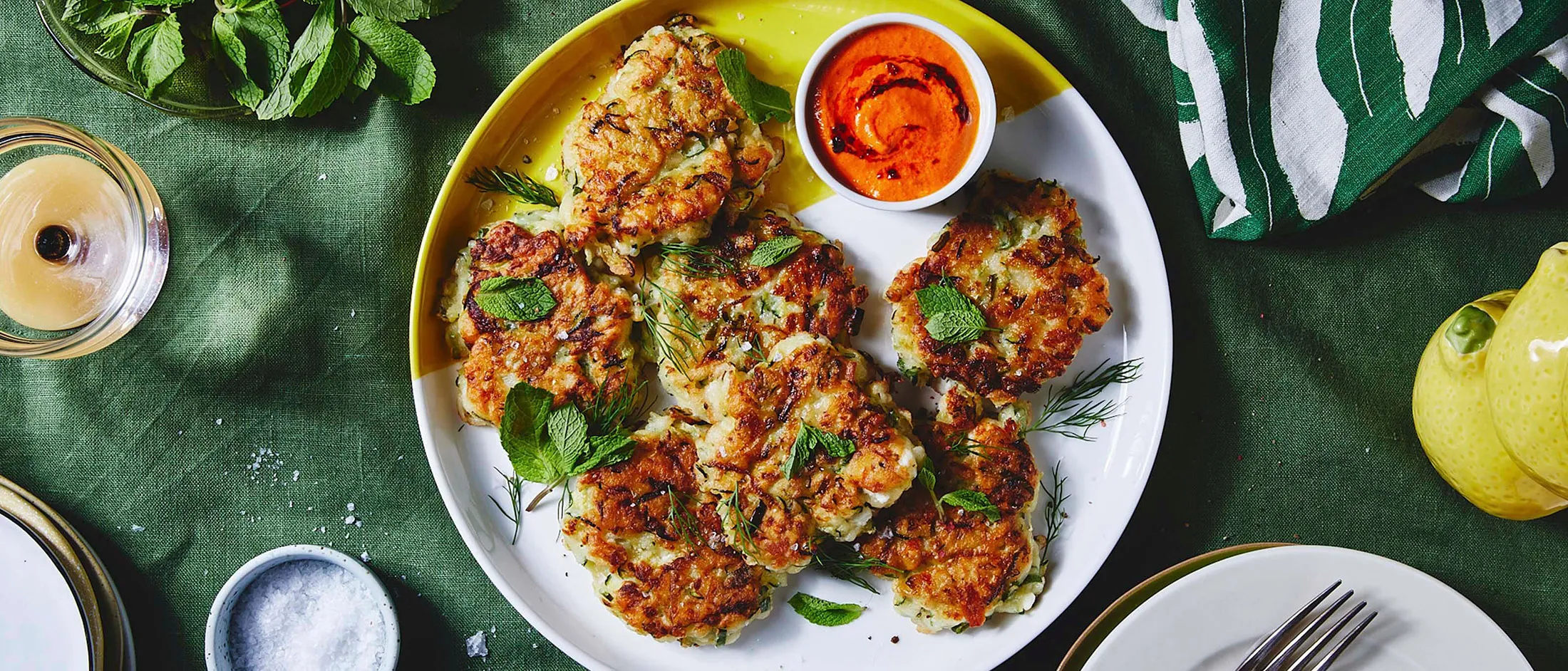A yellow and white plate with zucchini fritters and red dip on a green table cloth surrounded by colorful dinner table dishes, like a fork, a napkin, plates and a glas filled with water.