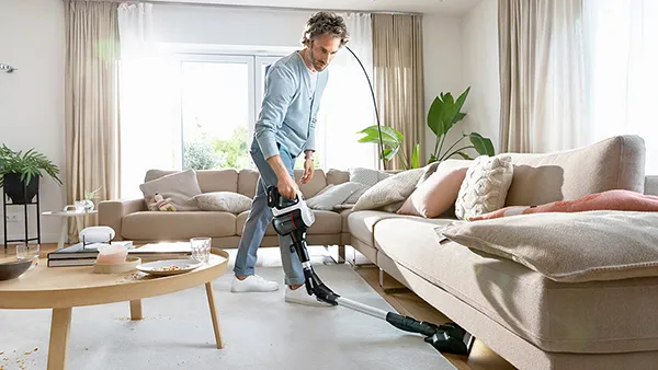 Ein Mann in legerer Kleidung saugt einen hellen Teppich in einem lichtdurchfluteten Wohnzimmer, das mit Zimmerpflanzen, einem Holzcouchtisch und einer großen beigefarbenen Couch eingerichtet ist.