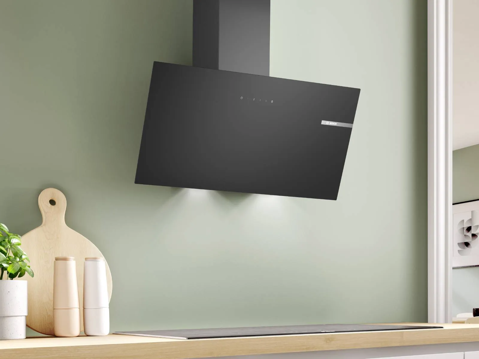 A sleek black Bosch angled extractor hood with integrated lighting is mounted on a sage green kitchen wall above a black induction hob and wooden countertop.