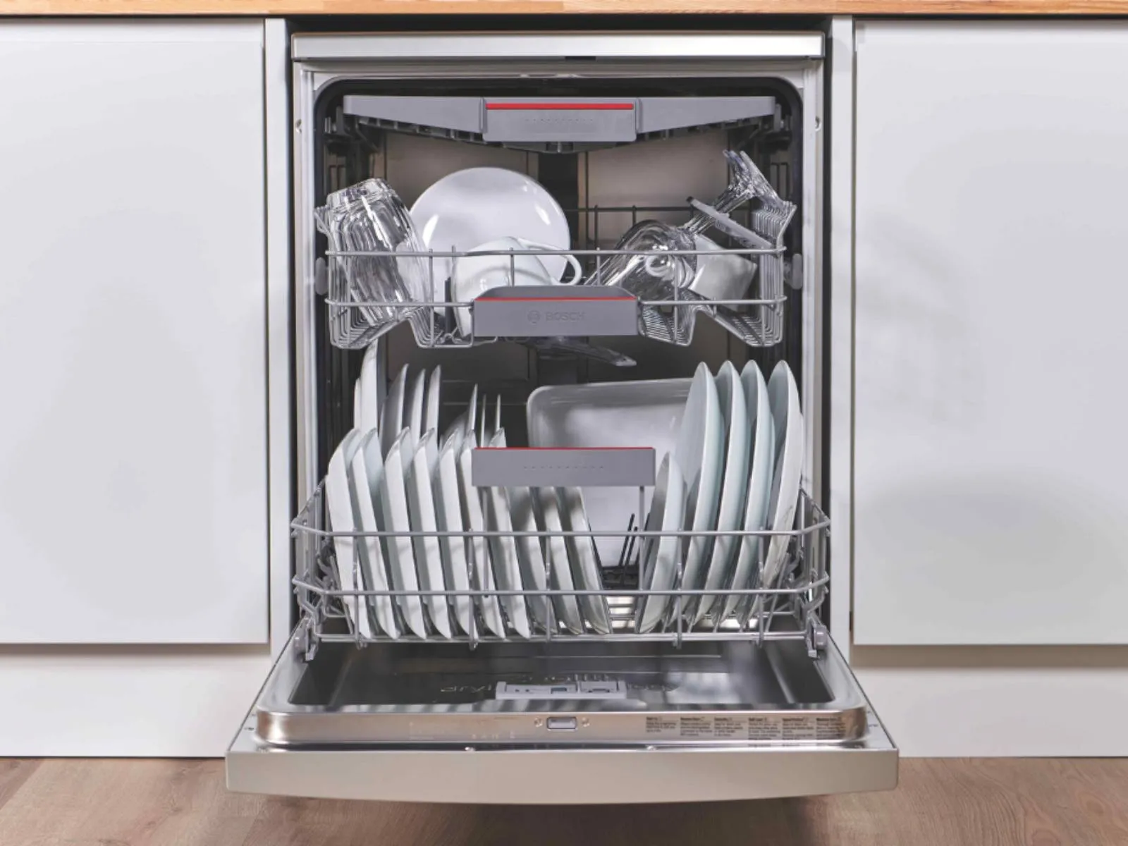 An open Bosch dishwasher filled with clean white plates, bowls, and clear glasses, integrated into a modern kitchen.