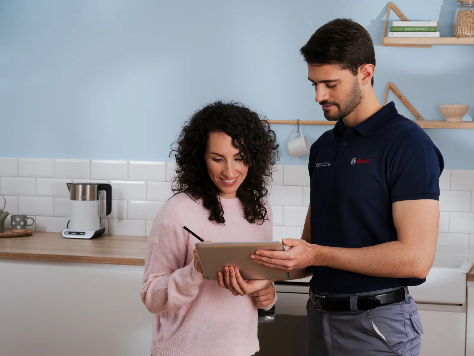 En Bosch-tekniker i handsker reparerer et apparat i et moderne køkken, mens en kvinde ser på. Værktøj ligger på bordet.