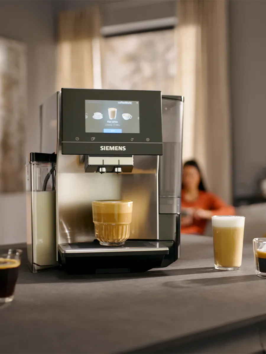 A Siemens coffee machine making a latte, surrounded by various coffee drinks and a plate of macarons, with a person relaxing in the background.