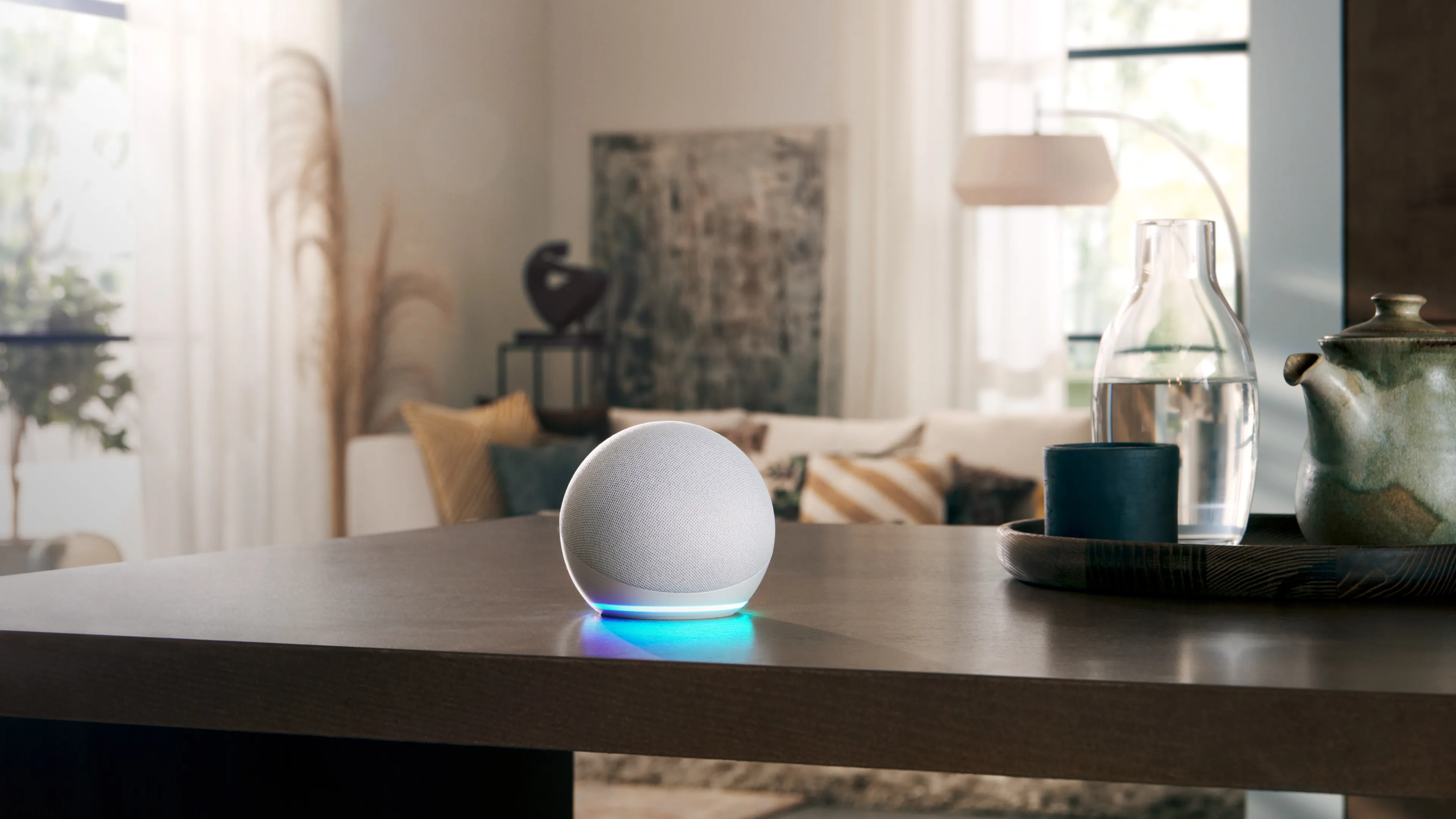 An Amazon Echo Dot smart speaker is sitting on a dark wooden table in a modern living room. Its blue light is illuminated, indicating it is active. In the blurred background, there is a comfortable-looking sofa, a floor lamp, and a tray with a glass carafe and a ceramic teapot.