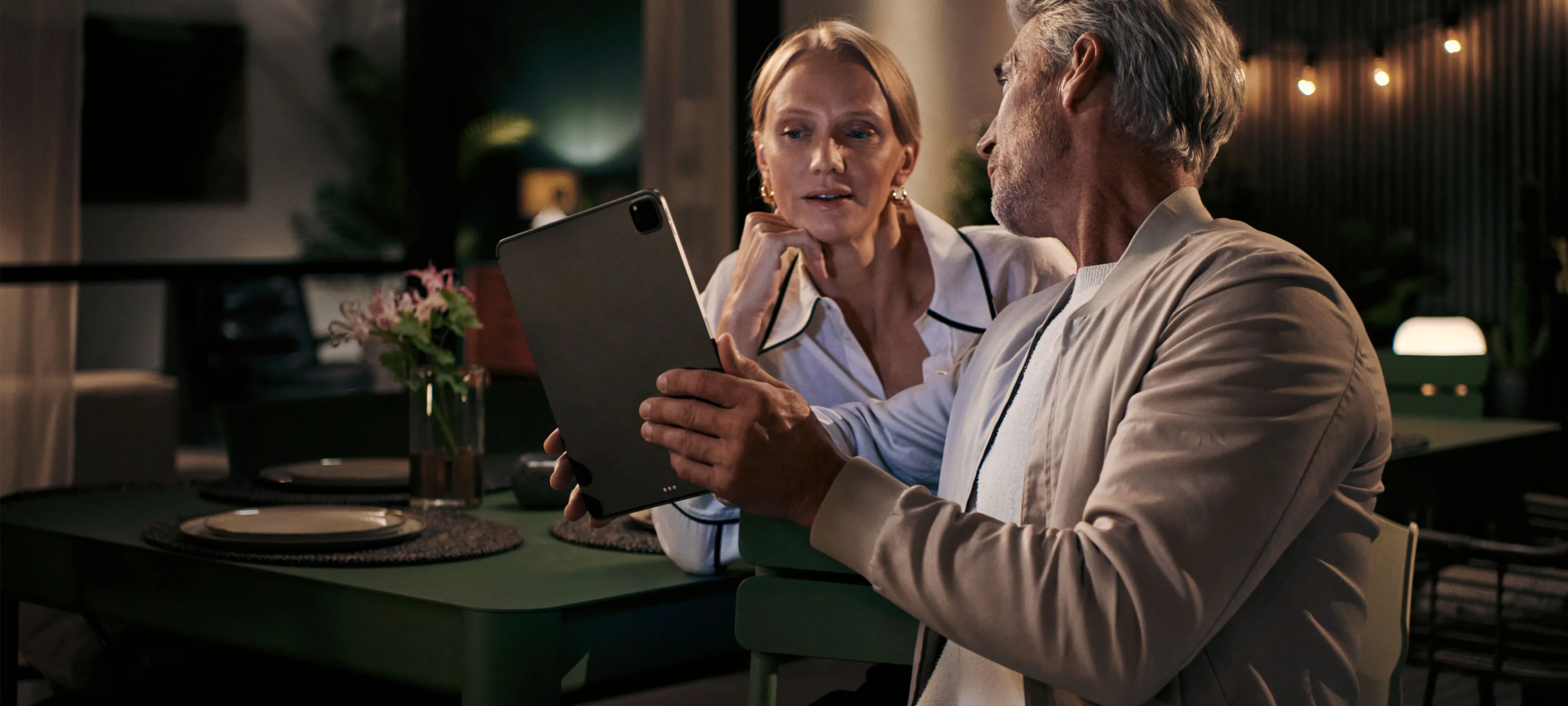 A mature man and woman are sitting at a dark green table on a patio at night, looking at a tablet that the man is holding. The woman is leaning in, engaged in what is on the screen. The scene is warmly lit by a table lamp and string lights in the background, creating an intimate and cozy atmosphere.