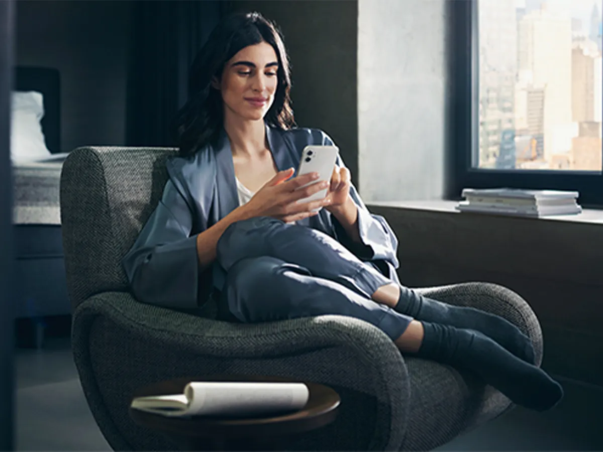 A woman relaxes in a stylish grey armchair in a modern apartment, smiling as she looks at her smartphone. She is comfortably dressed in blue loungewear, with a city view visible through the window beside her.