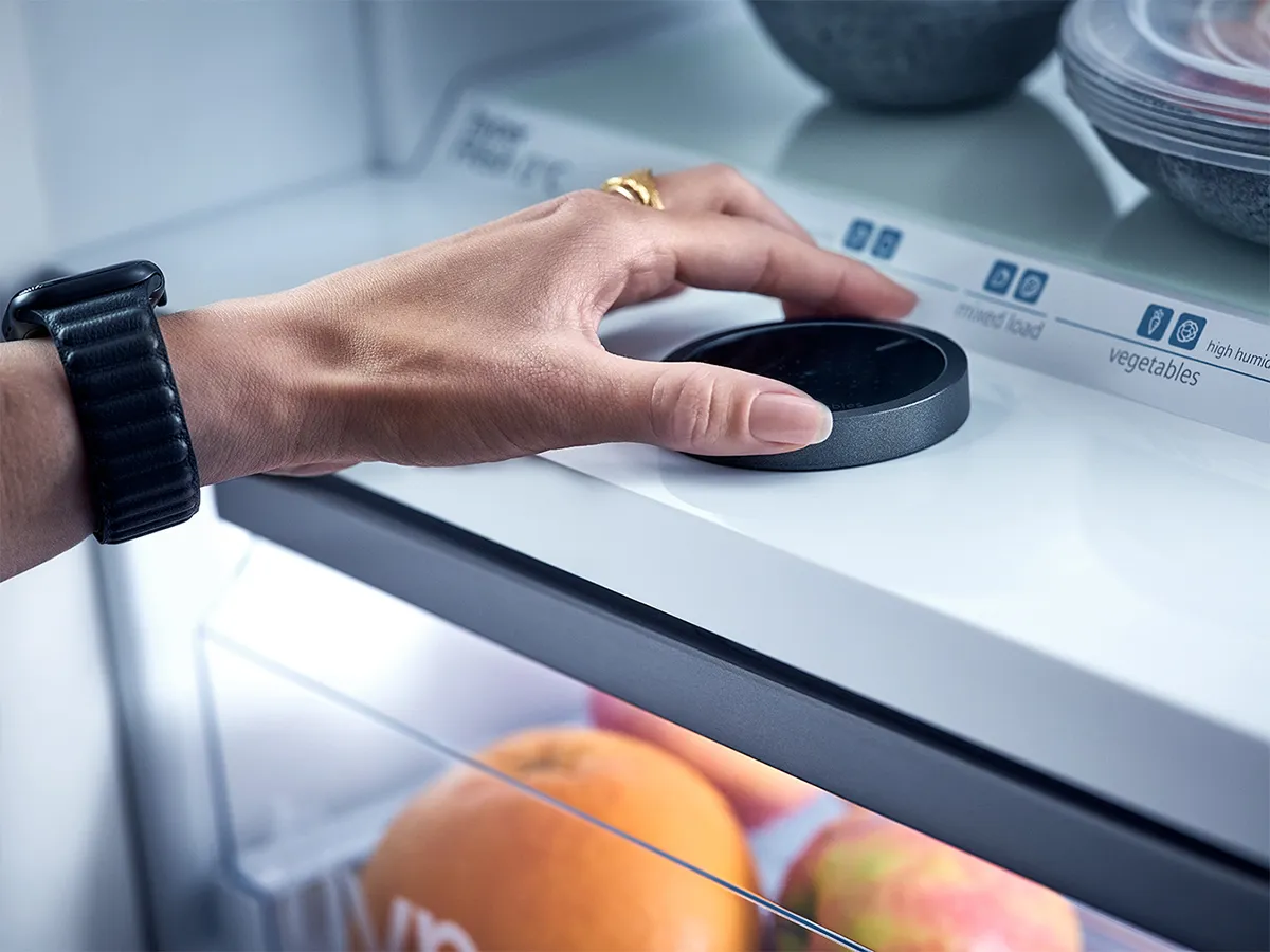 A person's hand is shown adjusting the circular dial on a Siemens hyperFresh refrigerator drawer to control the humidity levels for different food types, such as "vegetables" or "mixed load." Below the control, fresh oranges and apples are visible inside the crisper drawer.