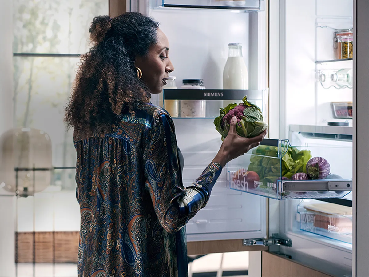 A woman in a paisley-patterned blouse is placing a fresh head of purple cauliflower into the "hyperFresh" drawer of a modern, built-in Siemens refrigerator.