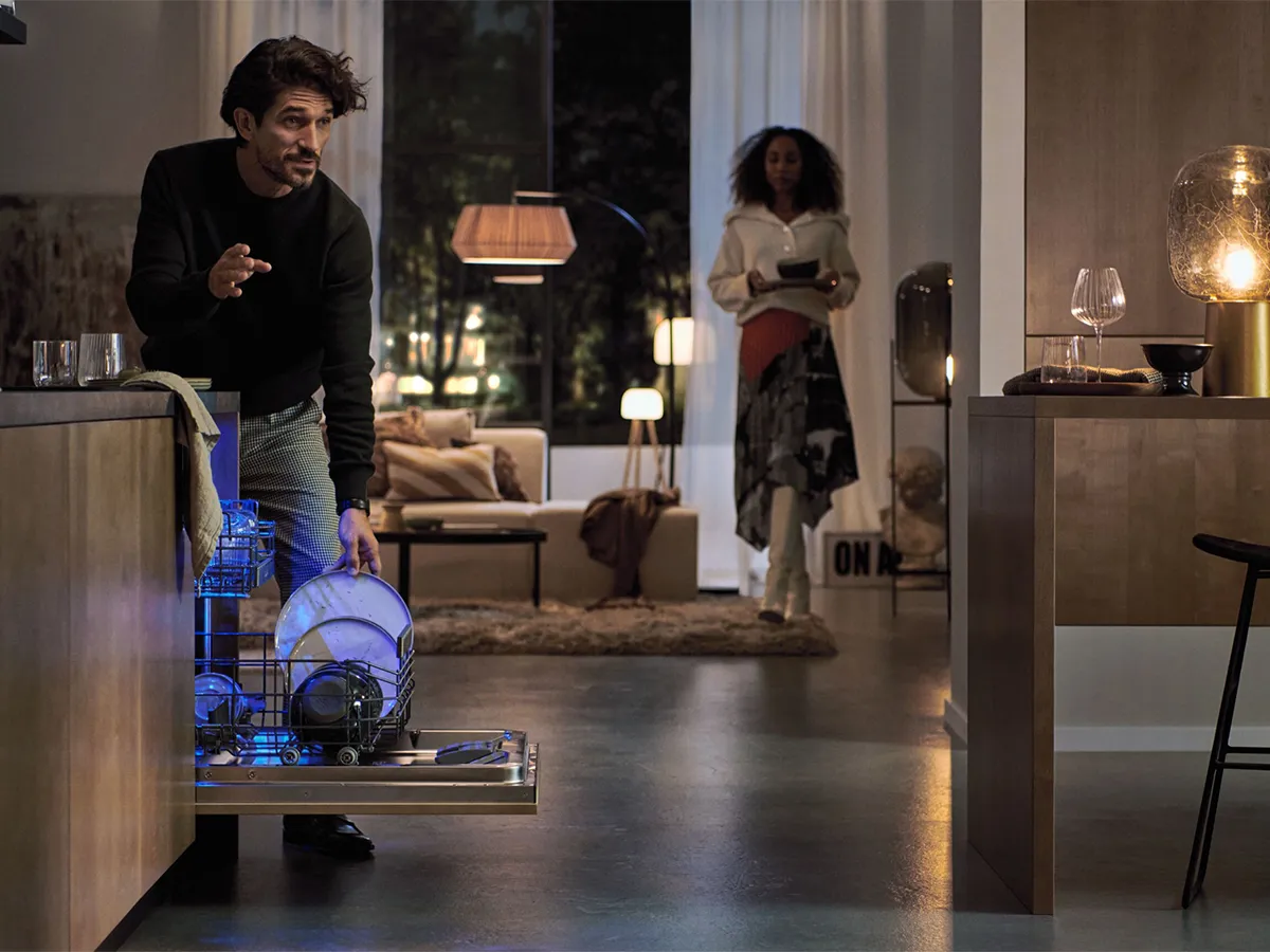 A man loading a blue-lit dishwasher in a modern open-plan kitchen and living room at night, with a woman in the background.
