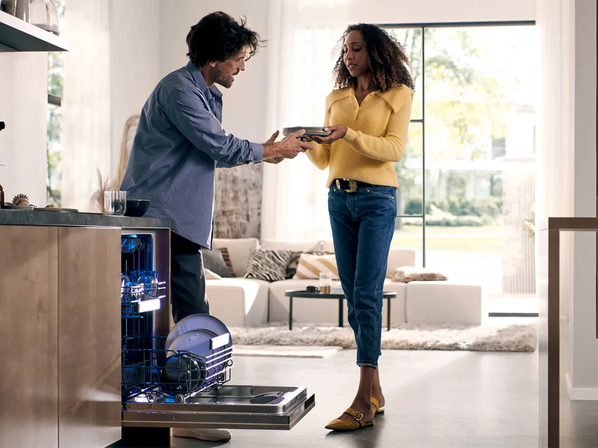 A man hands clean plates from a blue-lit dishwasher to a woman in a modern open-plan kitchen.