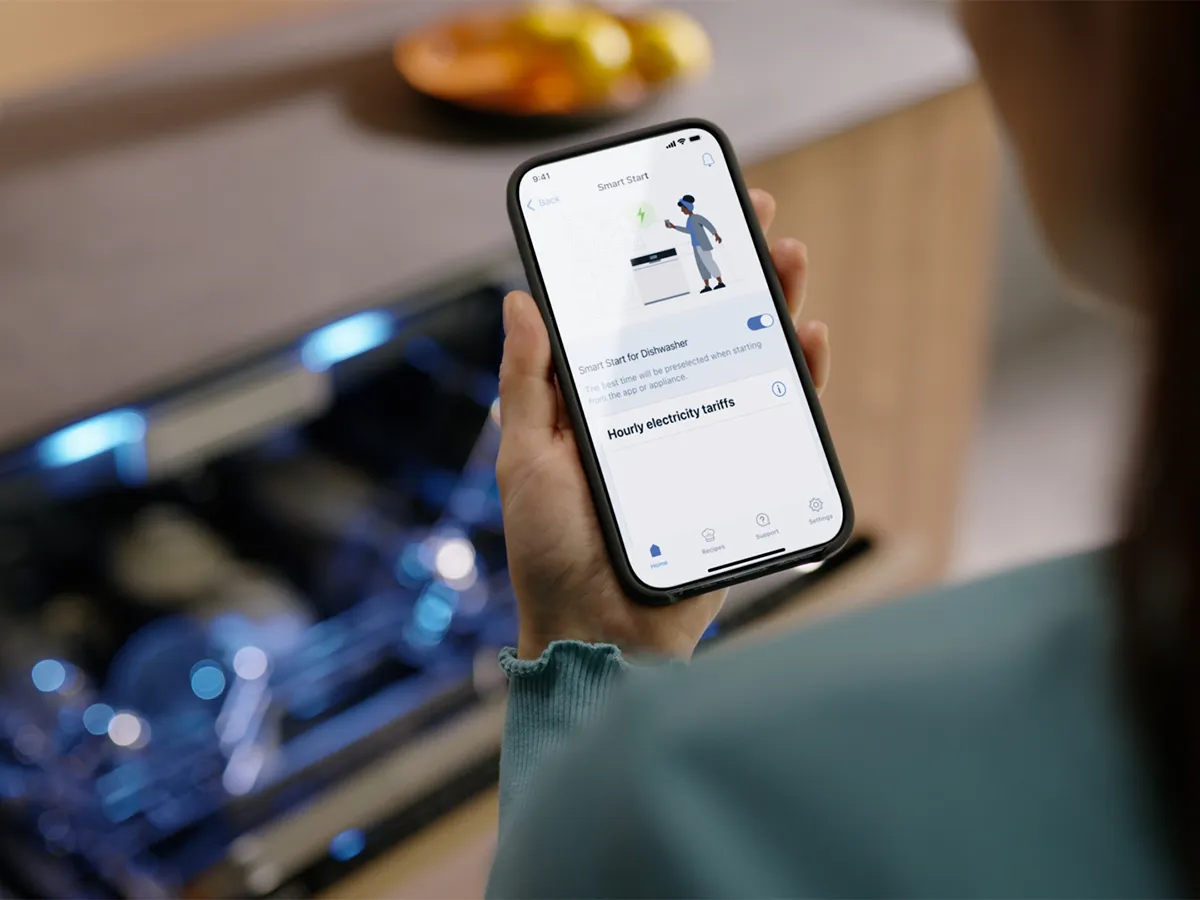 A person using a smartphone app to activate 'Smart Start' for a dishwasher, with the blue-lit dishwasher open in the background.