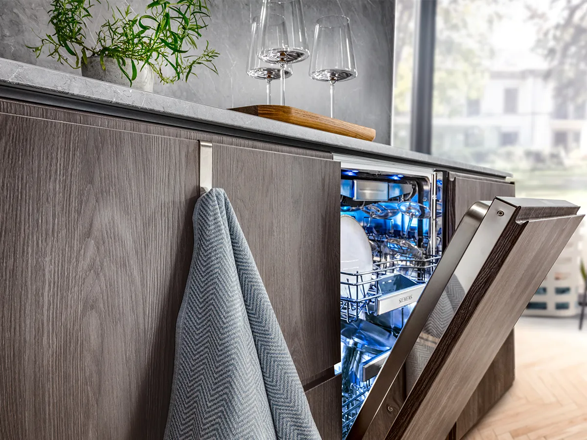 An open, integrated dishwasher with a blue interior light and clean dishes, in a modern kitchen with wooden cabinets.