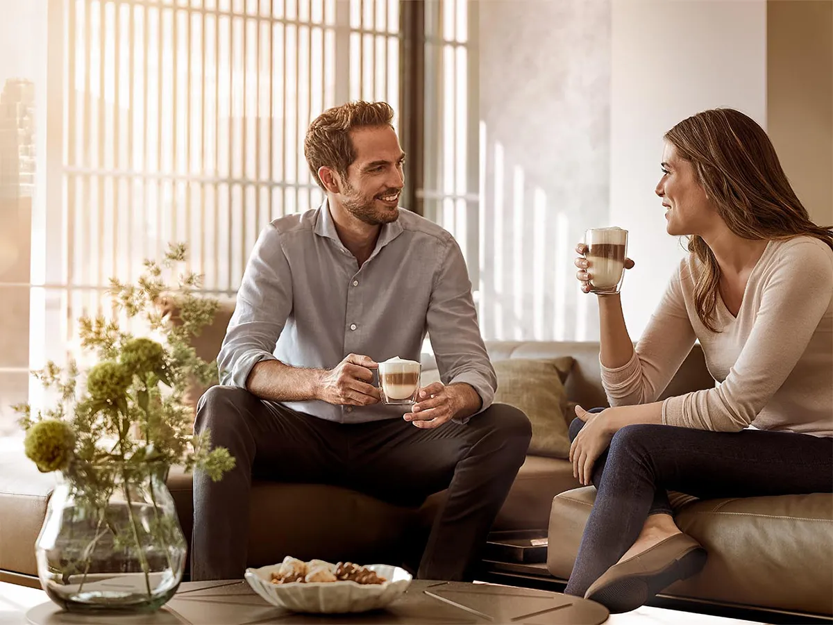 A smiling man and woman sit together on a comfortable sofa in a bright living room, enjoying a conversation over cups of cappuccino.