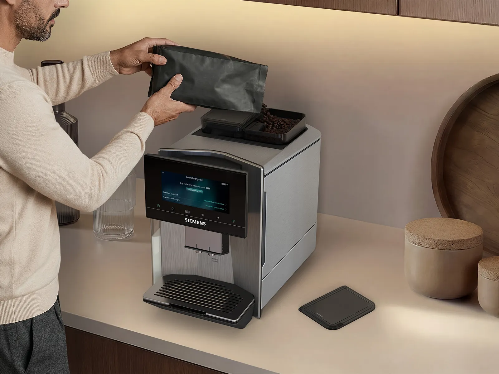 Siemens coffee machine on a light countertop; a person pours coffee beans into the top bean compartment, with textured glass containers nearby against a warm, neutral kitchen backdrop.