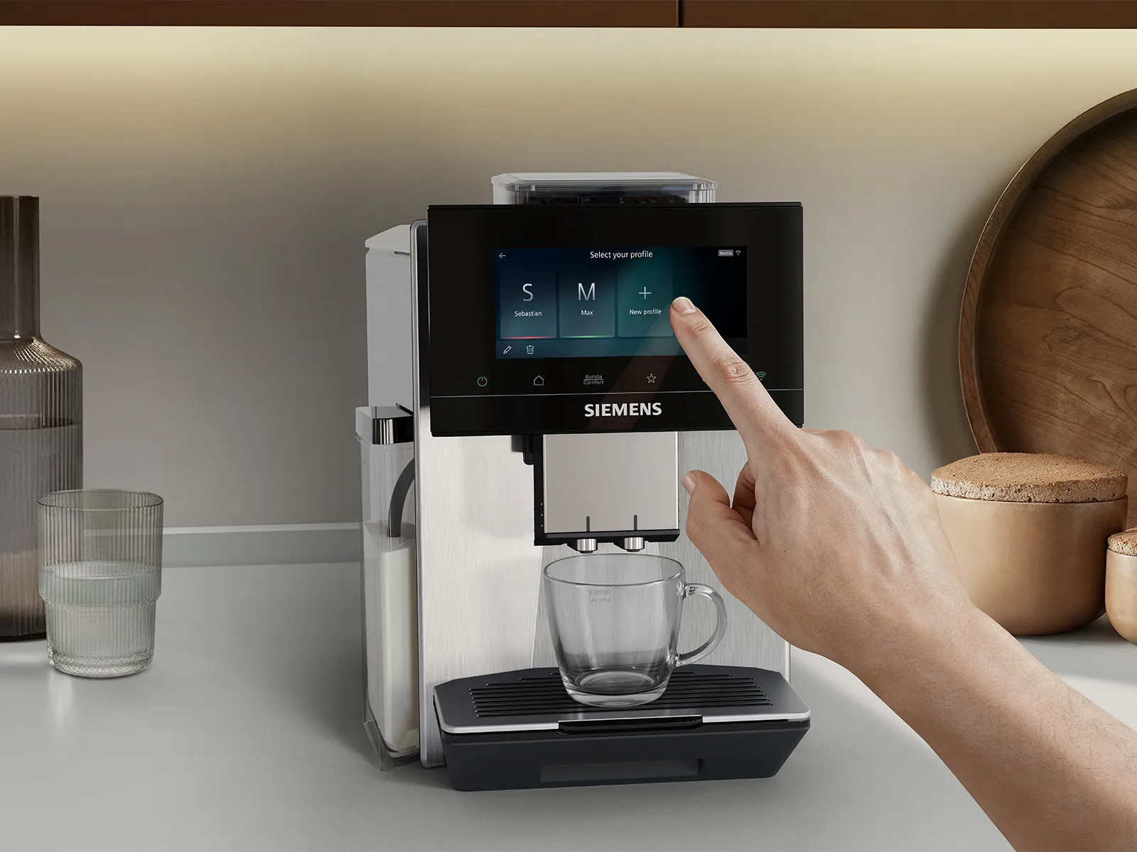 Hand reaching to a Siemens coffee machine touchscreen to select user profiles like Sebastian or Max; clear glass on the drip tray and a visible milk container, with wooden bowls and plate in a warm, well-lit kitchen.