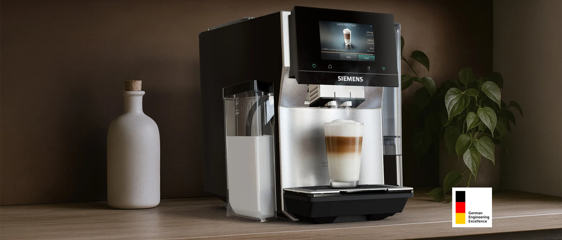 Siemens coffee machine with attached milk container on a wooden countertop with a layered latte macchiato; small beige vase with wooden lid and small plant in vase. “German Engineering Excellence” logo visible.