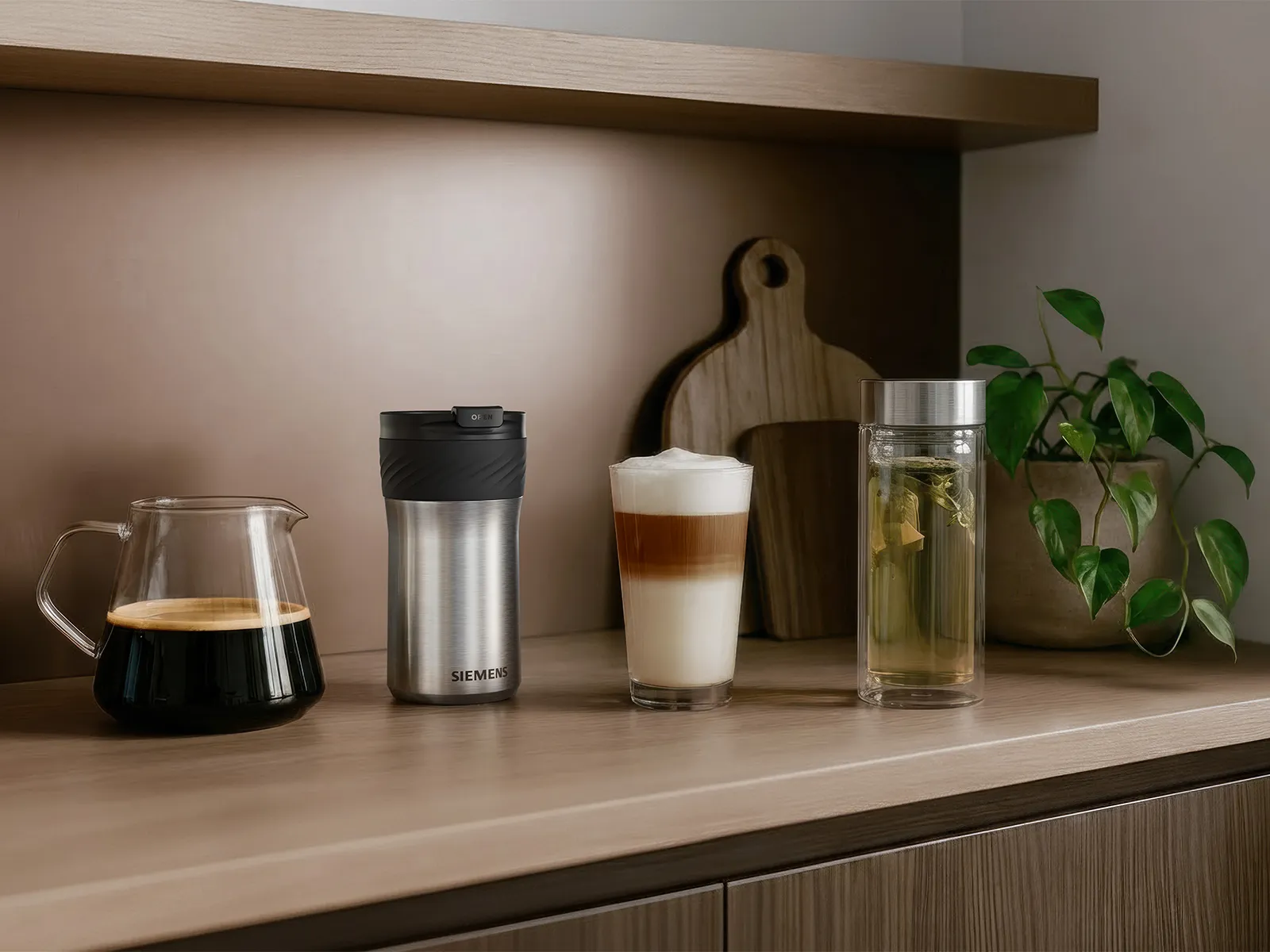 Kitchen countertop with Siemens travel mug, glass coffee carafe of dark coffee, layered latte in a clear glass, and a tall glass of tea with leaves and lemon; two wooden cutting boards and potted plant in background.