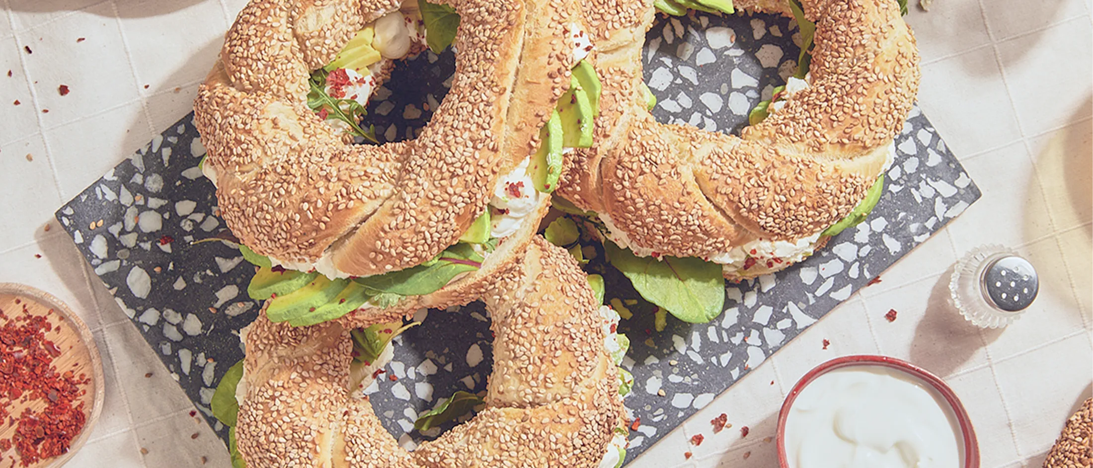 Drei Simits liegen auf einem schwarz-weißen Steinbrett auf einer weißen Tischdecke, umgeben von Geschirr.