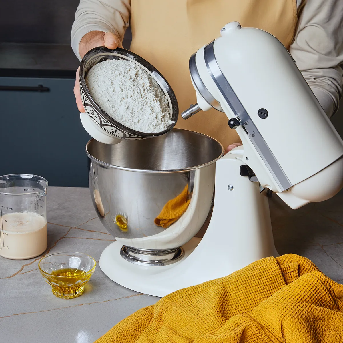 Eine Person gibt Mehl aus einer Schüssel in die Rührschüssel einer Küchenmaschine.
