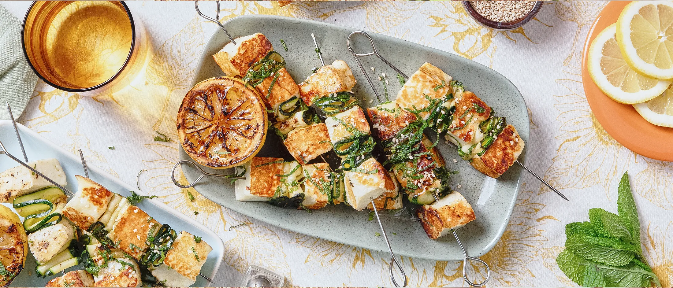 Gebratene Halloumi-Stücke auf Metallgrillspießen liegen auf einem blauen rechteckigen Teller, umgeben von anderen Tellern und Geschirr.