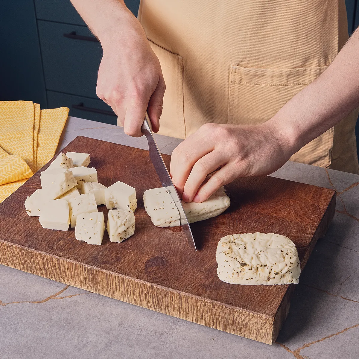 A person in an apron slices a block of cheese on a wooden cutting board, surrounded by cubed cheese and a yellow cloth.