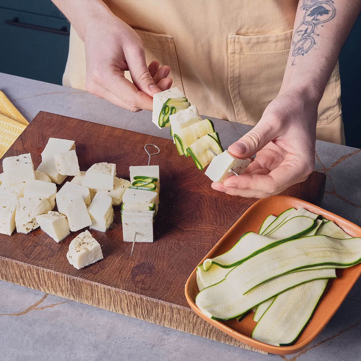 A person is assembling skewers with cubed cheese and zucchini slices on a wooden cutting board.