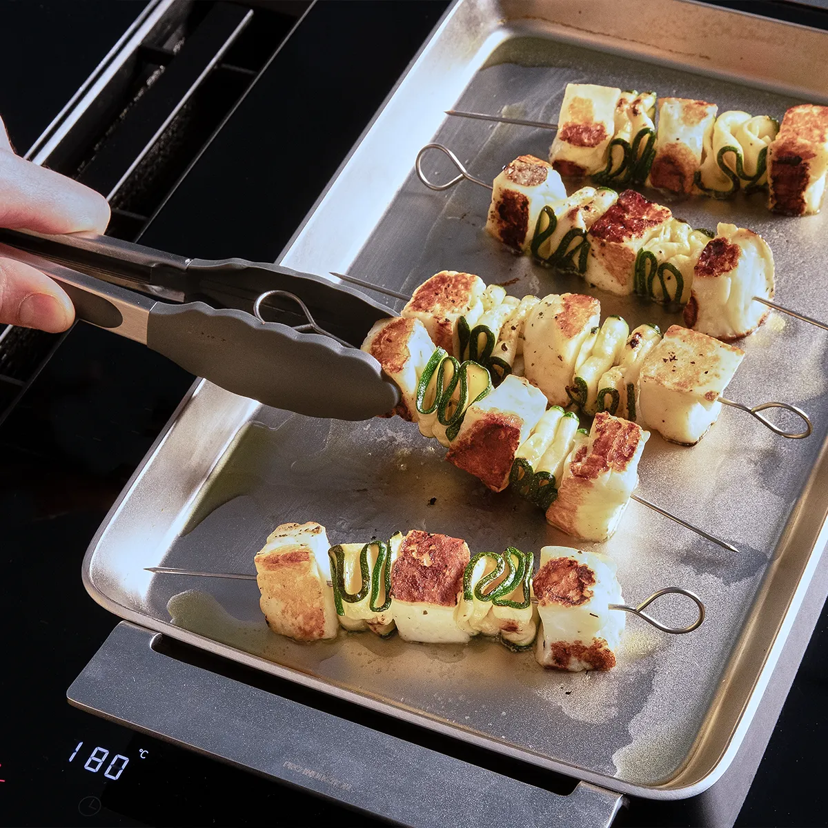 Grilled skewers of paneer and green peppers on a metal tray, with tongs lifting one skewer off an electric stovetop.