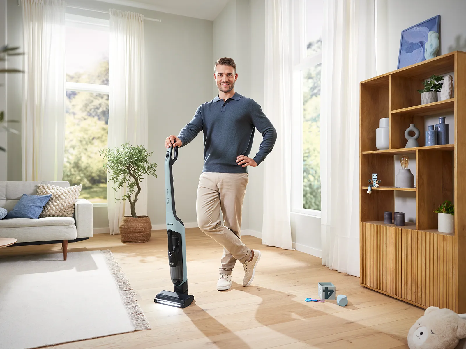 Мъж стои щастливо до прахосмукачка Flexxo Серия 4 в светла всекидневна. Той се обляга с едната си ръка на прахосмукачката.