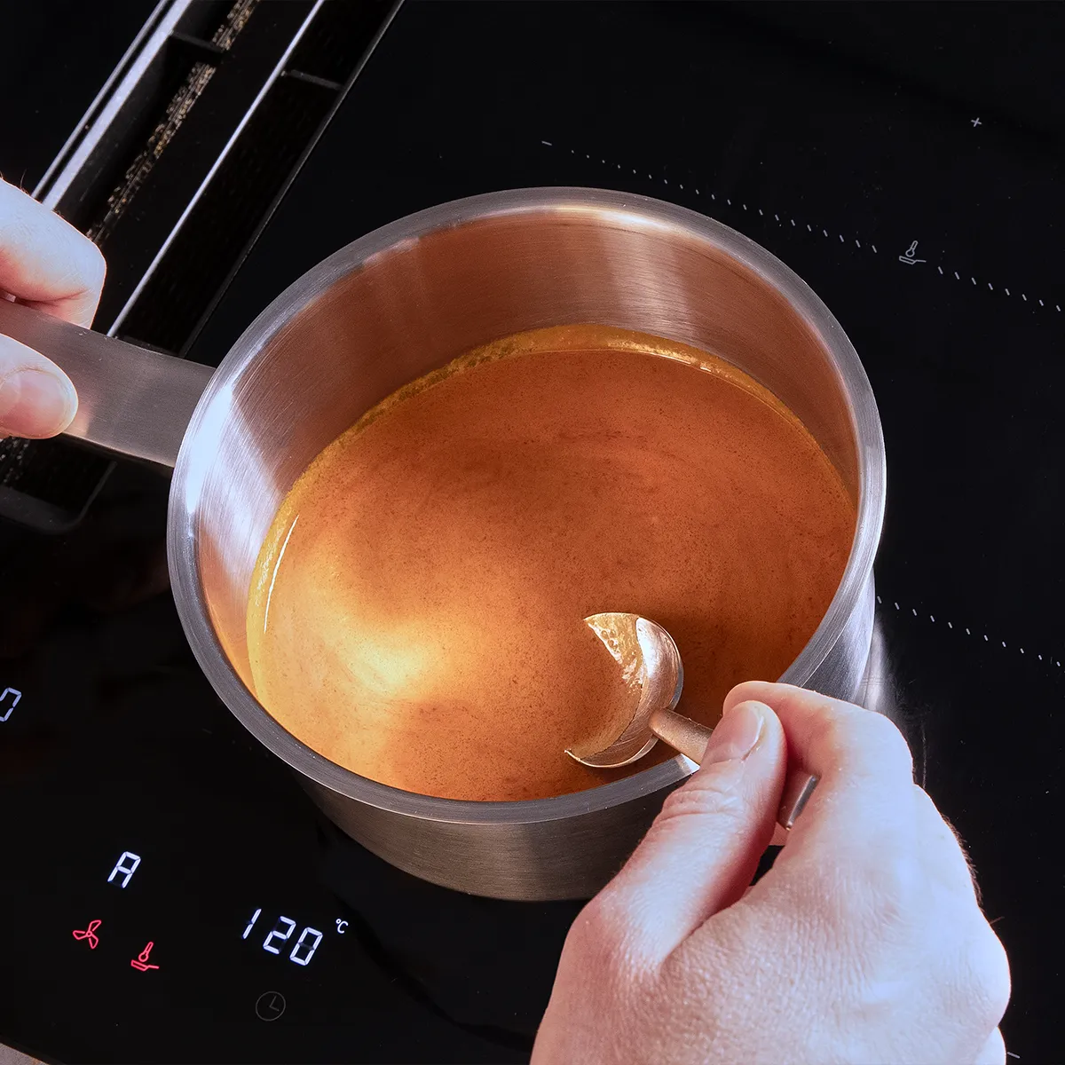 A person stirs a rich, brown sauce in a stainless steel pot on an induction cooktop, with a temperature display of 120°C.
