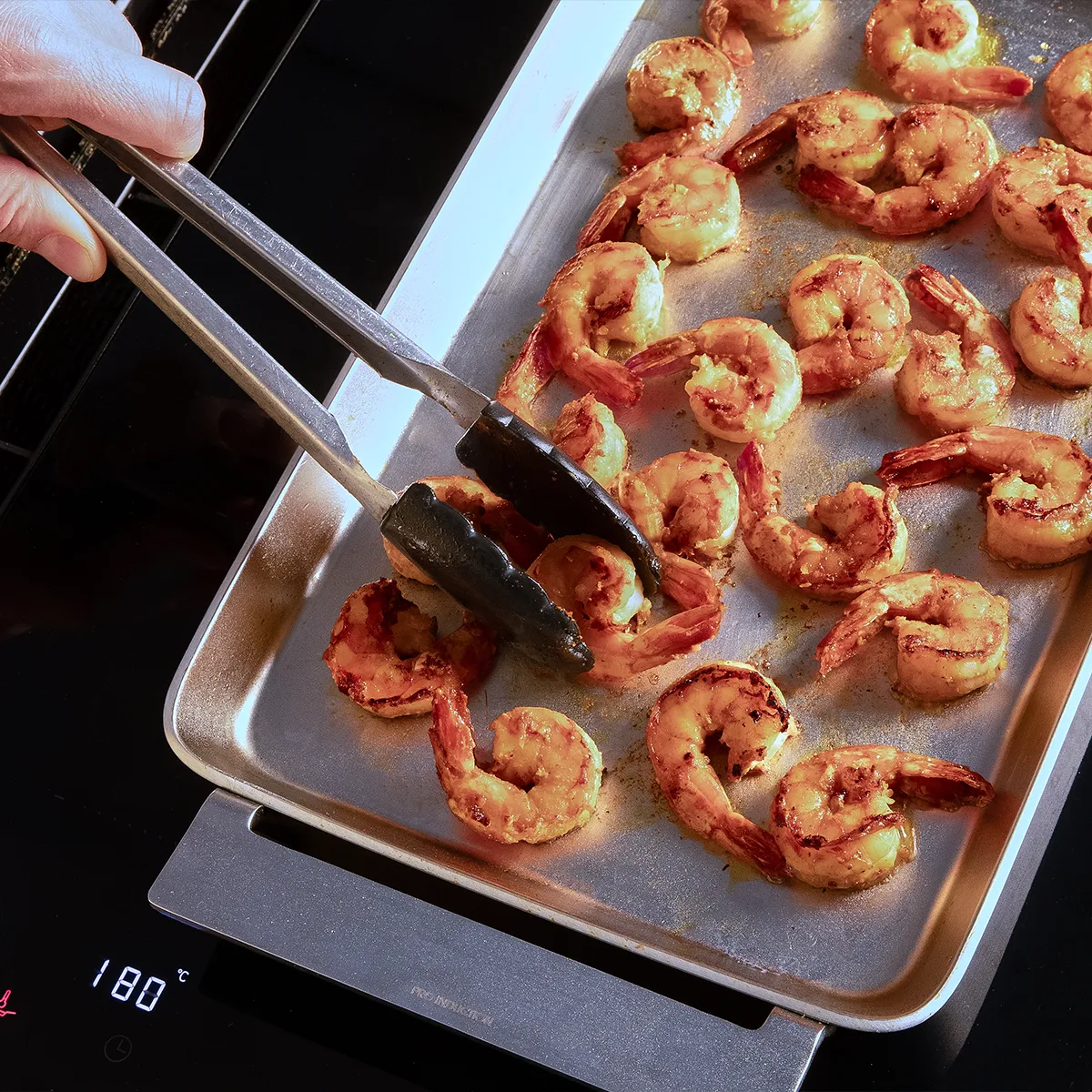 A hand using tongs picks up shrimp from a baking tray, with multiple perfectly cooked shrimp arranged on a metal tray.