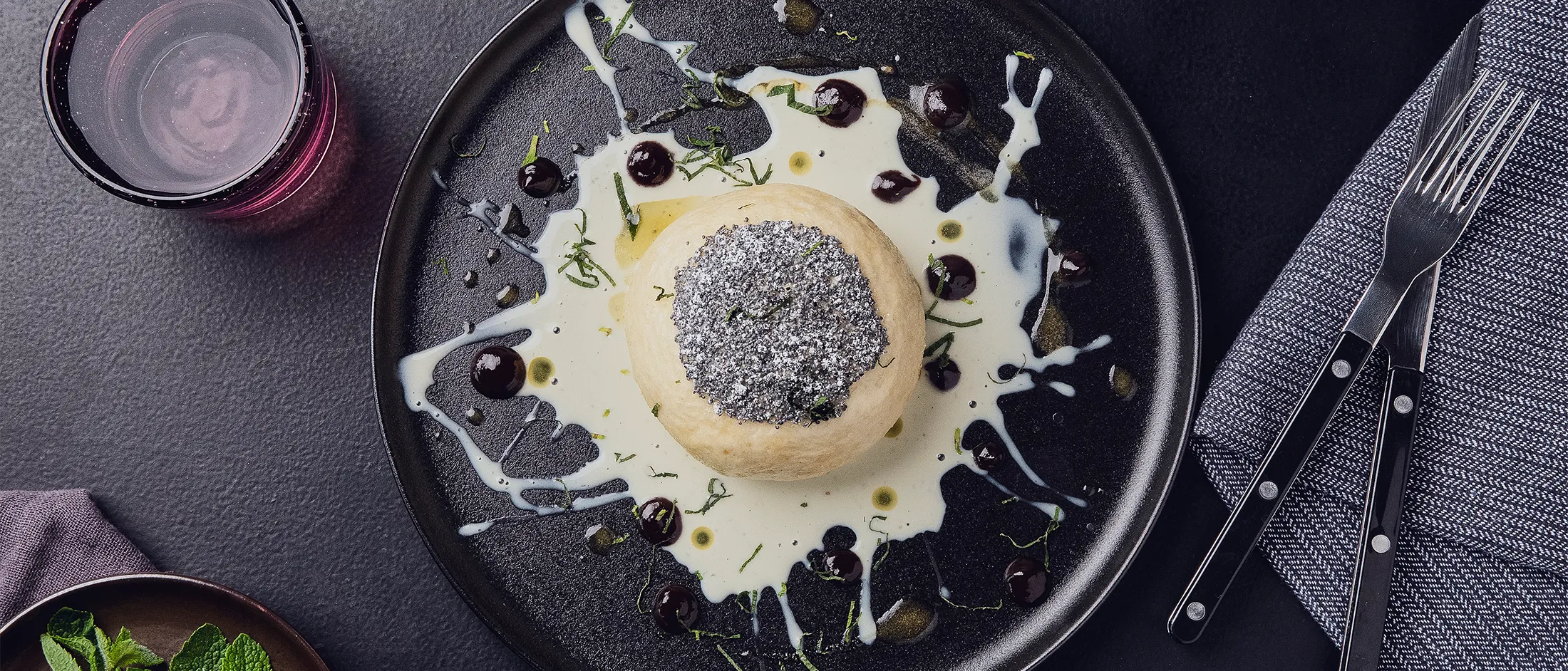 Einzelner Hefekloß, mit Mohn und Puderzucker bestreut, daneben Vanillesauce und Pflaumensirup, auf einem schwarzen Teller.