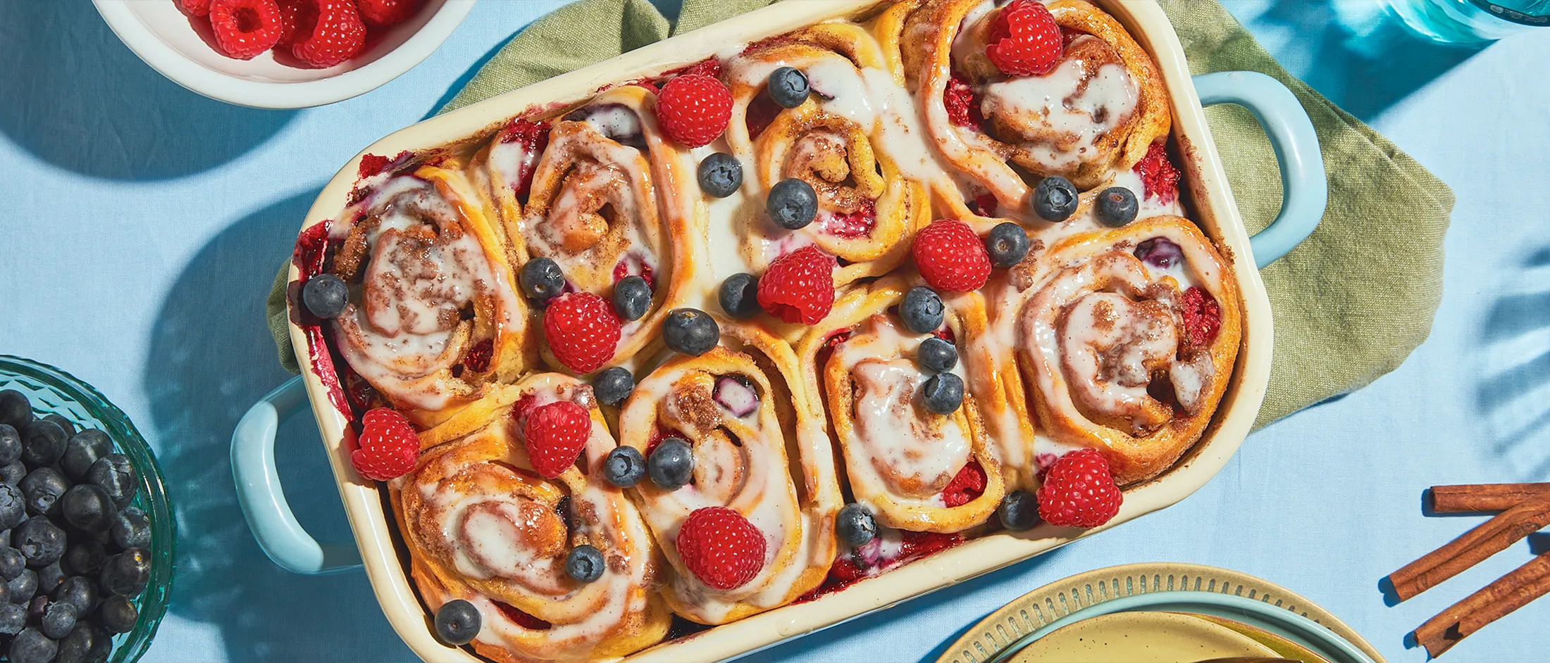 Gebackene Zimtbrötchen mit frischen Beeren und weißem Frosting in einer rechteckigen Backform stehen auf grünen und blauen Tischdecken, umgeben von Geschirr.