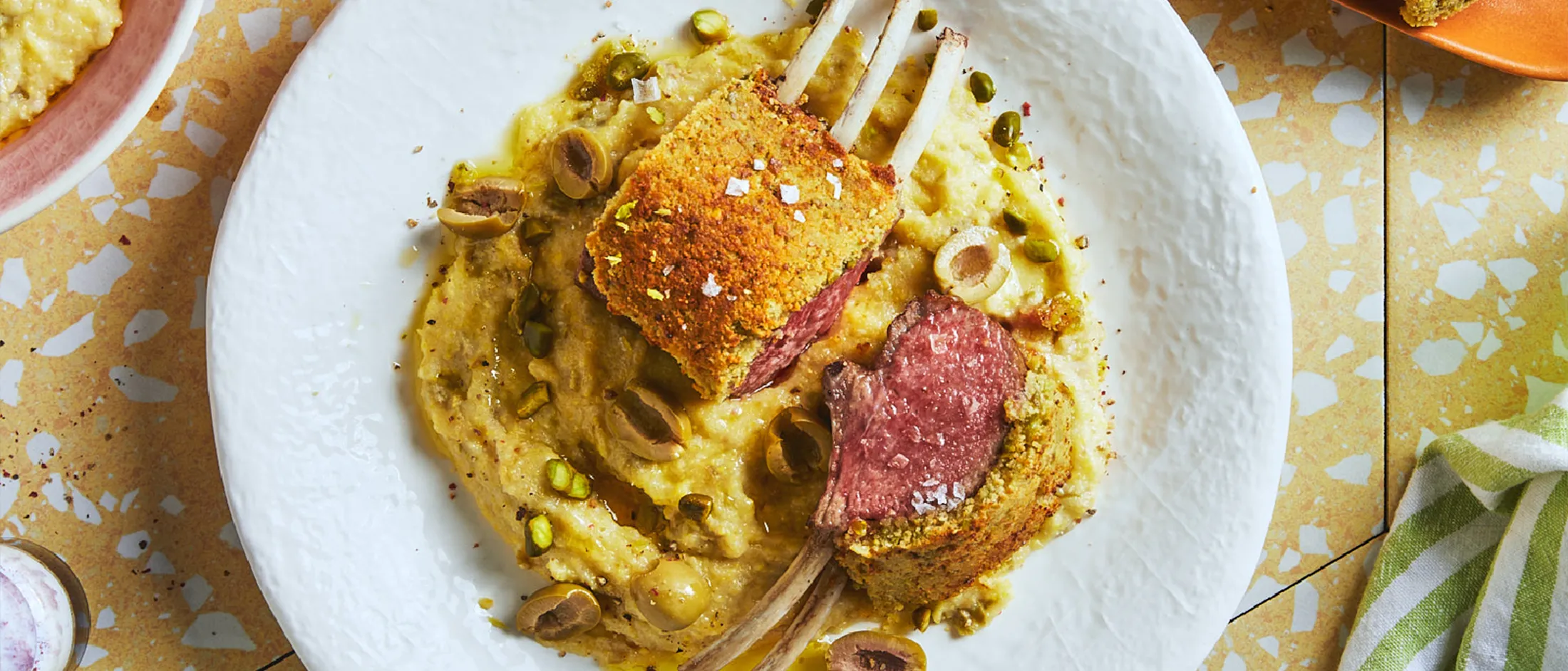 Auf gelben Keramikfliesen steht ein weißer Teller mit Lammkarree auf Polenta, umgeben von anderem Geschirr.
