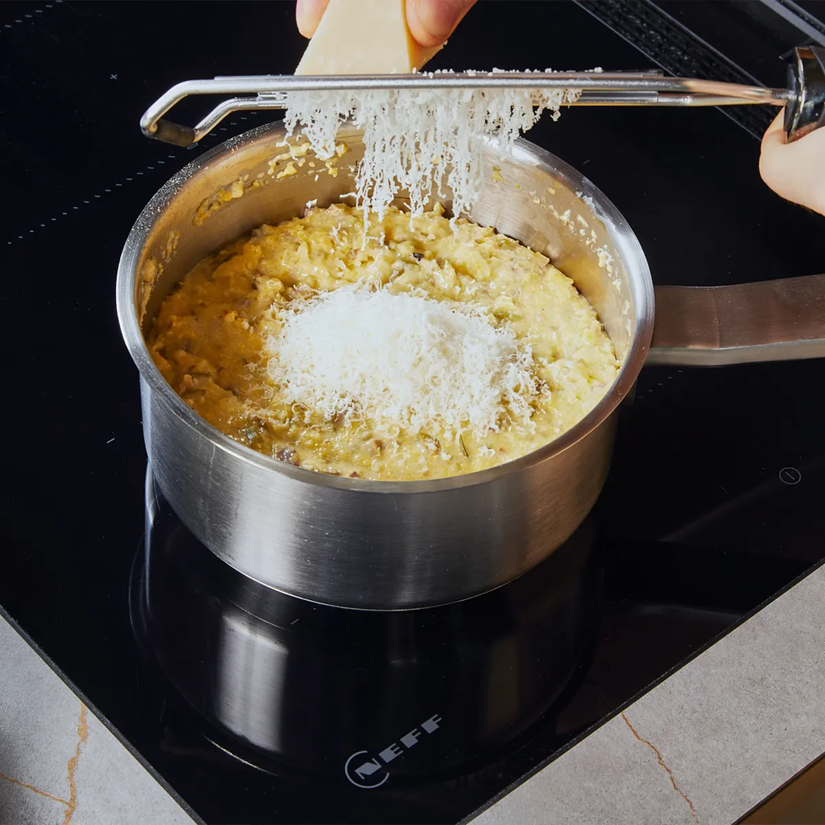 Grating parmesan into polenta within a silver pot on a stovetop.