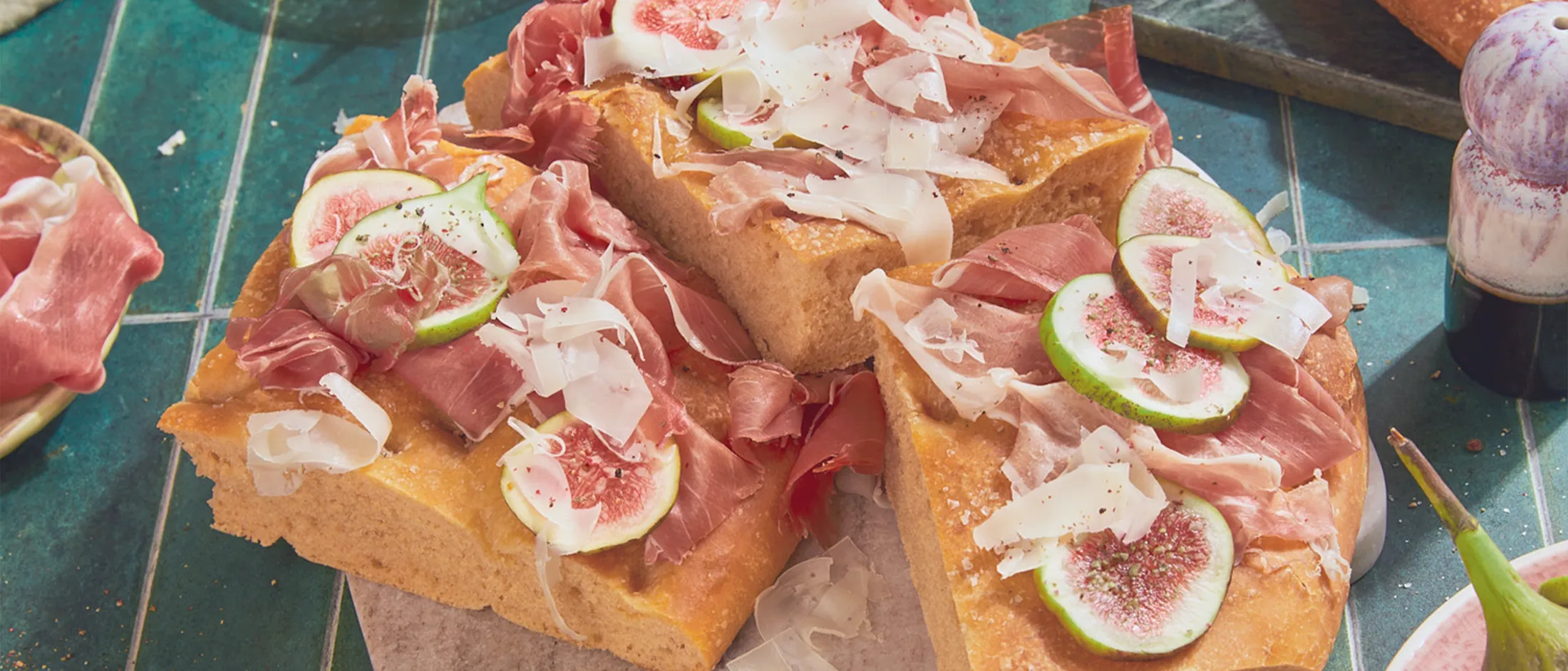 Tre pezzi di focaccia con prosciutto, formaggio e fette di fichi si trovano al centro dell'immagine su piastrelle da cucina blu e sono circondati da stoviglie.