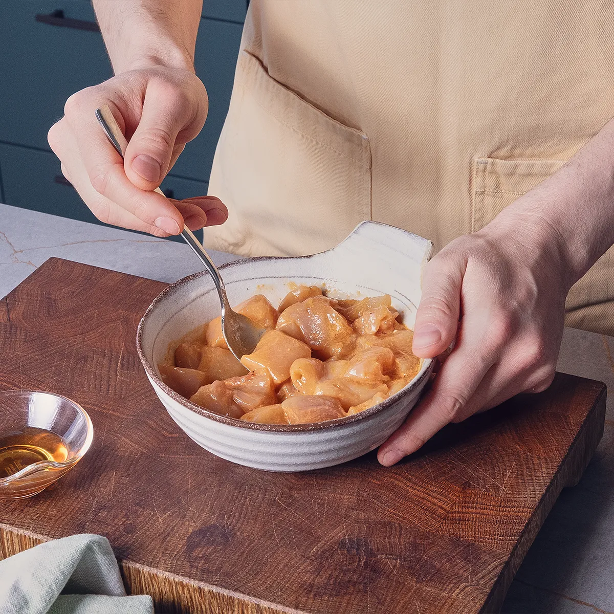 A person stirs marinated chicken pieces in a bowl on a wooden cutting board, with a small dish of sauce nearby.